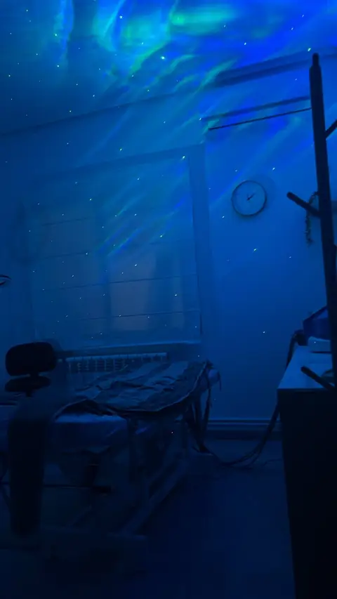 La cabina a oscuras, con una aurora azul y verde y puntitos de estrellas proyectados en el techo, el reloj y la camilla